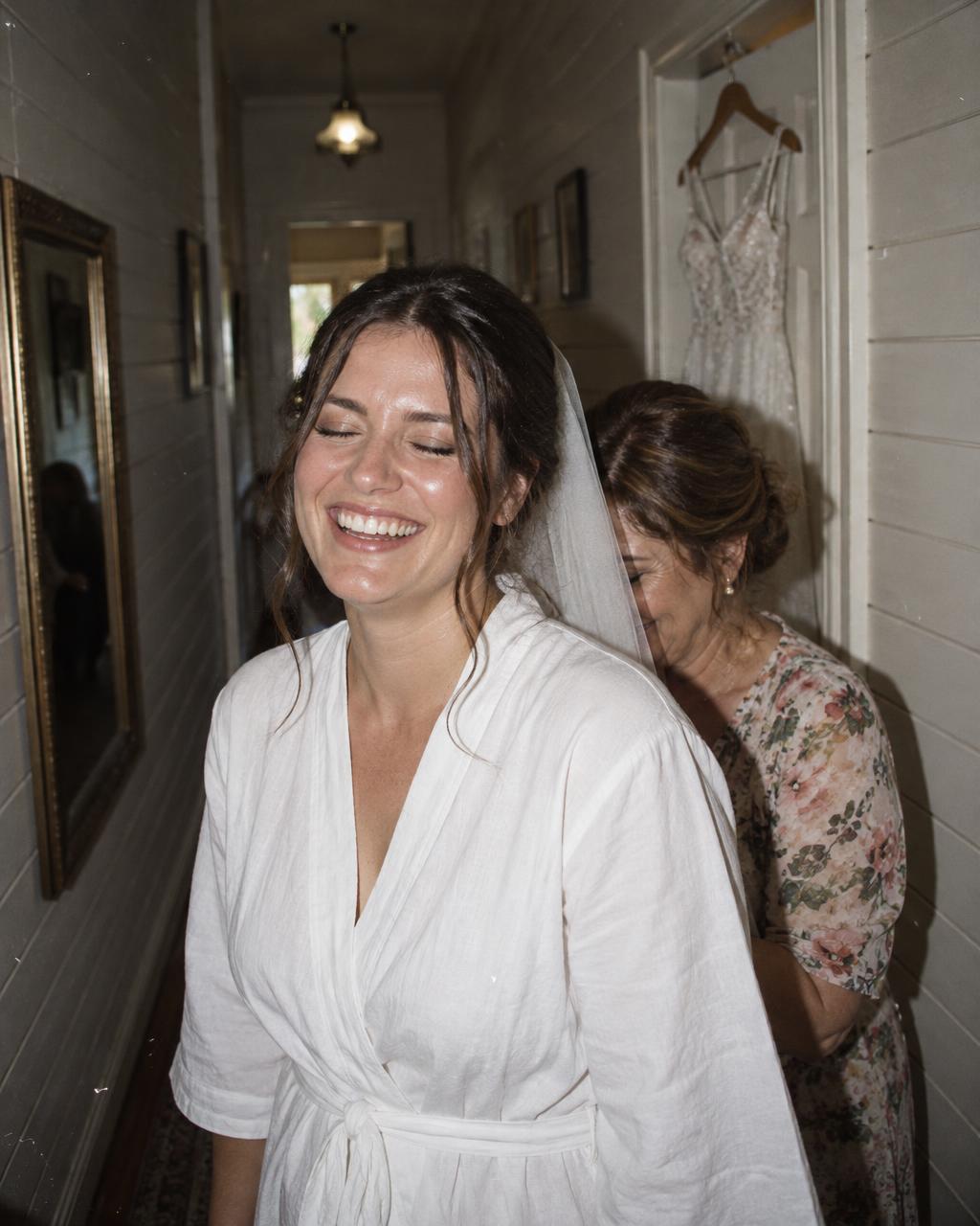 A bride laughing with her eyes closed as an older woman adjusts her veil in a timber-panelled hallway, a lace gown hanging in the background