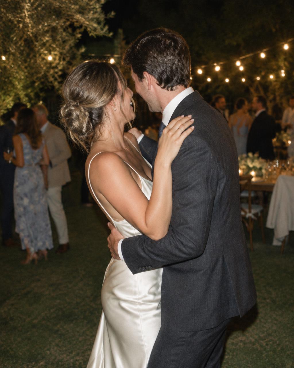 A bride in an ivory silk gown and her partner in a dark suit laughing as they dance close together at an evening garden reception under string lights